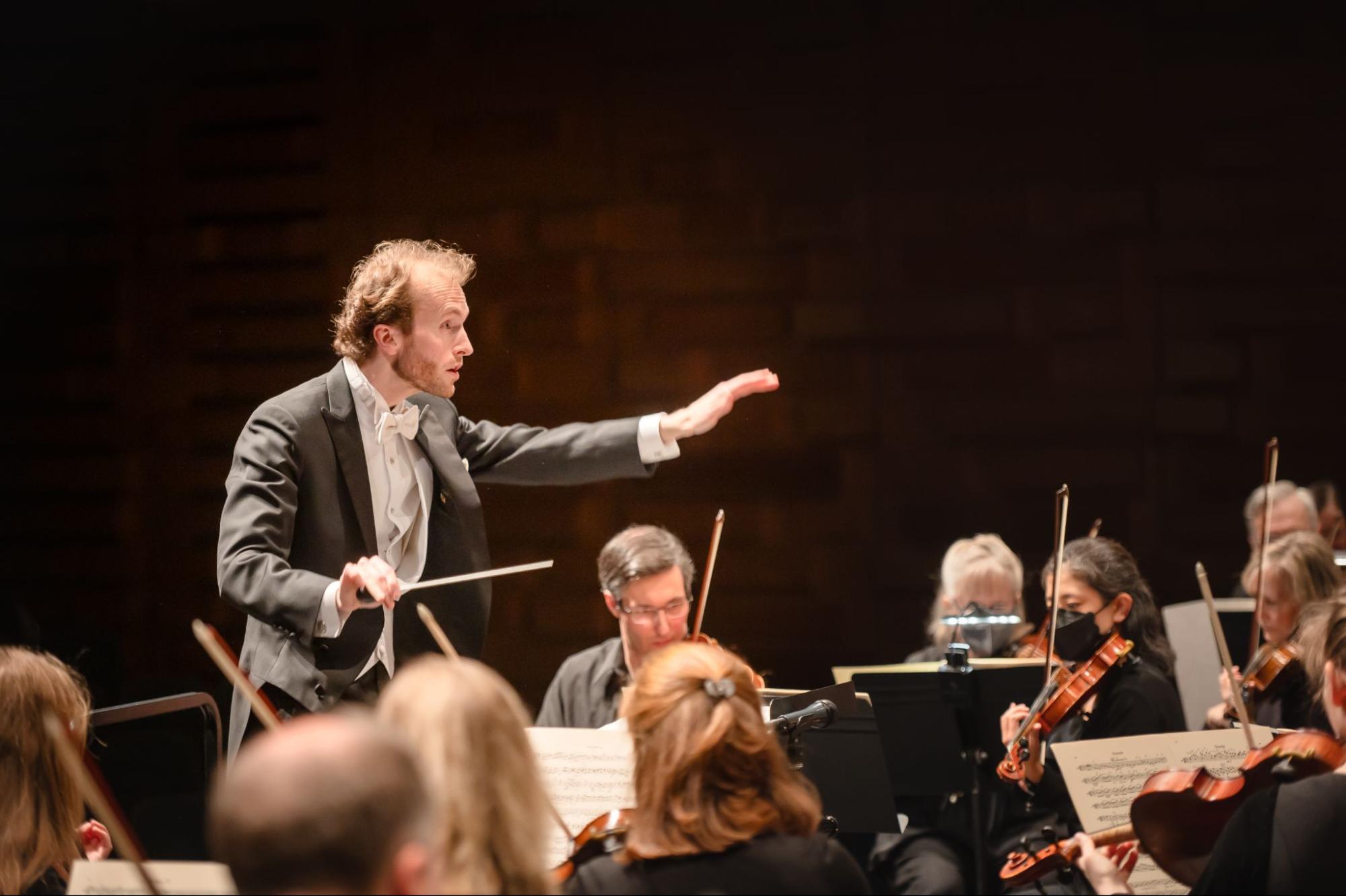 A conductor holds out his arms, one holding a baton, while conducting an orchestra. Several violinists are visible including June Spector, who is wearing a face mask.