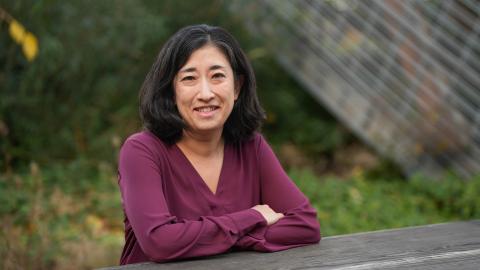 June Spector leans forward on a wood surface outside with arms crossed, wearing a maroon blouse. Behind her is a glass and steel sculpture.