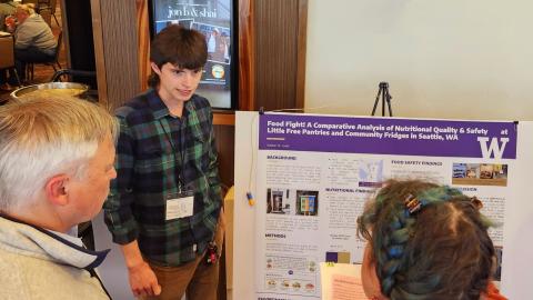 Kaidan St Louis wears a green and blue plaid shirt and stands in front of a research poster, explaining it to two observers.